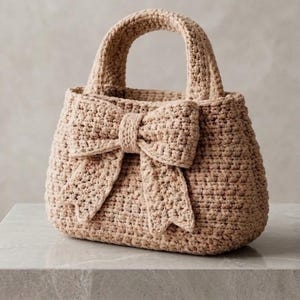 Puede incluir: Bolso de mano beige tejido a ganchillo con un gran lazo en la parte delantera. El bolso tiene forma redondeada y un asa resistente. La textura del ganchillo es visible, lo que sugiere un artículo hecho a mano. El bolso está sobre una superficie clara.