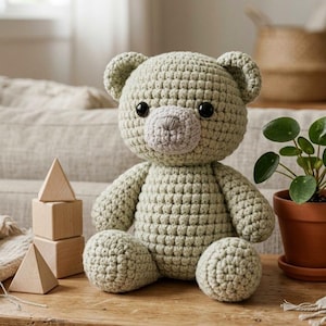 Pode incluir: Um urso de pelúcia de crochê feito à mão em um tom verde suave, com um focinho cinza claro e olhos pretos. O urso está sentado em uma superfície de madeira, com blocos de madeira e uma planta em vaso por perto. Um brinquedo artesanal e aconchegante.