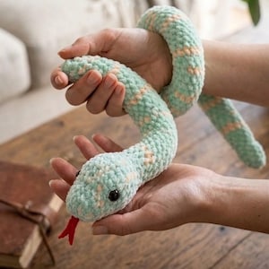 May include: A crocheted snake toy in mint green and peach. The snake has black button eyes and a red felt tongue. The soft toy is held in open hands, highlighting its texture and design. A charming, handmade item.