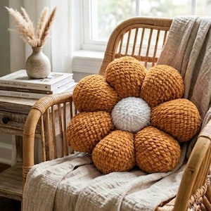 Peut inclure: Un coussin décoratif en forme de fleur, avec un centre blanc et des pétales brun doré, repose sur une couverture beige drapée sur une chaise en osier. La texture tricotée du coussin ajoute un élément décoratif et confortable. Une petite table de chevet et un vase sont visibles en arrière-plan.