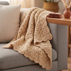 Peut inclure: Une couverture beige au crochet avec un bord festonné est drapée sur un canapé gris. La texture de la couverture est visible, avec un motif répétitif de petits points ouverts. Un coussin beige repose sur le canapé, et une table en bois avec des livres et un vase est à l'arrière-plan.