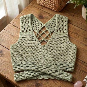 May include: A sage green crochet top with a crisscross back design. The sleeveless top features a V-neck and a wrap-around style at the waist. The crochet pattern includes openwork details, creating a textured and airy look. The top is displayed on a wooden surface.