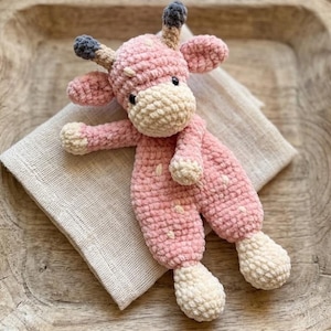 Peut inclure: Une girafe en crochet rose faite à la main avec des taches blanches, un museau crème et des cornes grises. L'animal en peluche est posé sur un tissu en lin de couleur neutre.