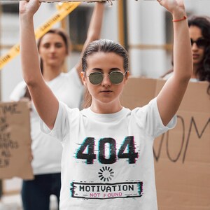 Könnte beinhalten: Ein weißes Kurzarm-T-Shirt mit einer schwarzen Grafik, die den Text 404 und ein Lade-Symbol über den Worten MOTIVATION NOT FOUND zeigt. Der Text und das Symbol weisen einen digitalen Glitch-Effekt mit roten und blauen Farbverschiebungen auf.