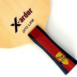 May include: A light-colored ping pong paddle with the text "X-ardor pro Line" in black and red. The handle is red and black with a gold and yellow emblem. The paddle is designed for table tennis.