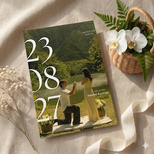 Puede incluir: Una invitación de boda con la fecha "23 08 27" en grandes números blancos. La imagen de la invitación muestra a una pareja, con el hombre arrodillado frente a la mujer. También hay una pequeña cesta de flores y una cinta.