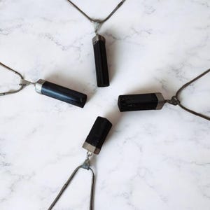 Può includere: Cinque collane con pendenti in cristallo di tormalina nera, decorate con dettagli in metallo argentato e catenine, sono disposte su una superficie di marmo bianco. I cristalli cilindrici sono di un nero intenso.