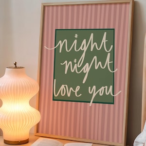 May include: A framed print with the words "night night love you" in white script on a green background, set against a pink striped backdrop. A glowing, ribbed glass lamp and an open book sit on a bedside table.
