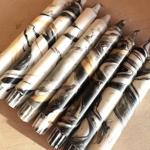 May include: A selection of marble-effect taper candles. The candles display a swirling pattern of black, white, and gold. The candles are arranged diagonally, highlighting their unique design and texture. These decorative candles are suitable for home decor.