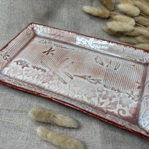 May include: Rectangular ceramic serving tray with a red rim and a textured, off-white surface. The tray features a floral and dragonfly design. Decorative dried plants are in the background.
