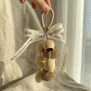 Op de afbeelding: Beige decoratieve hangende ornament met een lus en een wit lint. Op het lint staat "BEST WISHES FOR YOU". Het ornament heeft een cluster van houten en getextureerde bruine elementen.