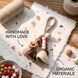 Peut inclure: Un ornement suspendu décoratif comprenant un cheval à bascule en bois brun, deux pendentifs en forme de cloche en brun foncé et crème, et des perles blanches et rouges sur un cordon beige tressé. Le texte indique HANDMADE WITH LOVE et ORGANIC MATERIALS.