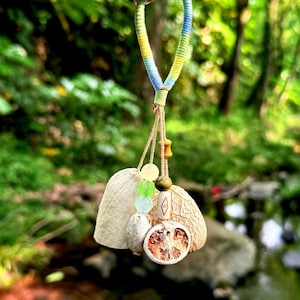 May include: A decorative hanging ornament featuring a braided loop in yellow, blue, and green. It includes natural elements like a dried seed pod, a sliced citrus piece, and several small round beads in green, yellow, and light blue colors.