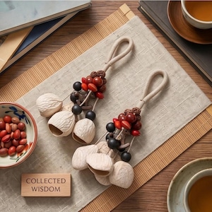 Peut inclure: Deux ornements décoratifs suspendus composés de grappes de coquilles de noix claires, de perles marron foncé et de petites graines rouges fixées à des boucles en corde beige. Un bloc en bois rectangulaire porte le texte COLLECTED WISDOM.