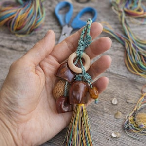 May include: A handcrafted keychain with a collection of polished brown wooden pieces, a wooden ring, and a vibrant tassel. The keychain is held in a hand, with blue scissors and colourful threads in the background.