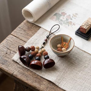 May include: A selection of handcrafted wooden beads and decorative items. The image shows a string of dark brown beads, a small ceramic bowl with decorative knots, and a wooden stamp on a wooden table. The items are arranged on a linen cloth.