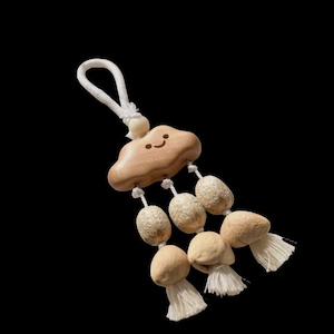 May include: A wooden cloud-shaped charm with a smiling face, connected to several beige, oval-shaped beads and tassels. The charm is suspended by a white rope loop against a black background.