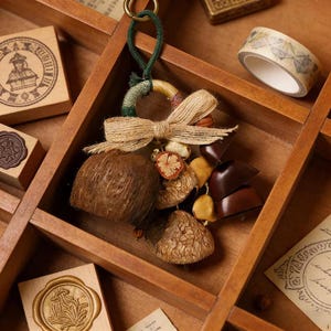 May include: A wooden tray filled with various decorative items. A handmade ornament with a jute bow and dried elements hangs in the center. Other items include wooden stamps, a roll of patterned tape, and dried seed pods. The color palette is warm, with browns and natural tones.