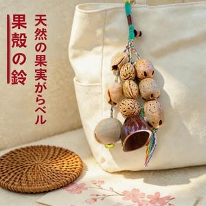 May include: A cluster of natural seed pods and a bell, strung together with colorful threads, hangs from a cream-colored tote bag. A woven coaster and a card with cherry blossom illustrations are in the foreground. Japanese text is visible.