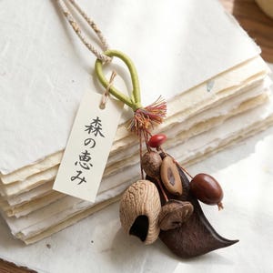 Peut inclure: Un pendentif décoratif composé d'une boucle en tissu vert clair, d'un cordon beige et d'un assortiment de graines et de gousses naturelles dans des tons marron et rouge. Une étiquette blanche rectangulaire porte le texte japonais 森の恵み.