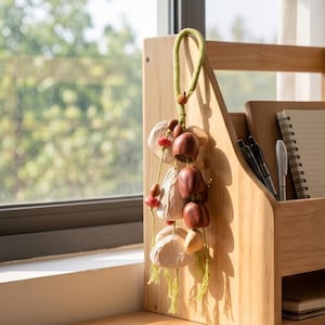 May include: A decorative hanging ornament featuring a light green braided cord loop, small brown and red wooden beads, and several smooth, rounded wooden shapes in shades of cream, tan, and dark brown, finished with light green fringe tassels.