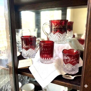 May include: A white ceramic mug with a black handle and the text "But First, Coffee" in a bold, black font. The mug is set against a blurred background, suggesting a cozy setting.