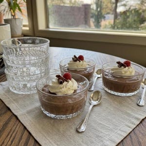 May include: A stack of clear glass dessert bowls with a textured pattern sits next to individual servings of chocolate mousse topped with whipped cream, raspberries, and chocolate shavings. Silver spoons are arranged around the desserts.