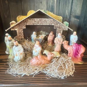 May include: A nativity scene set featuring a stable structure with a textured roof, containing figurines of figures in robes, a manger with an infant, two camels, and a sheep, all arranged on a bed of dried straw material.