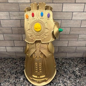 May include: A gold-colored gauntlet featuring a textured metallic finish and six embedded stones in orange, red, blue, purple, yellow, and green. The object displays intricate mechanical detailing across the knuckles, wrist, and forearm sections.