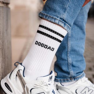Könnte beinhalten: Weiße Sportsocken mit zwei schwarzen Streifen und dem Wort "DOGDAD" in schwarzen Blockbuchstaben. Die Socken werden mit blauen Jeans und weißen Sportschuhen getragen.