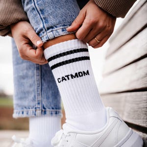 Könnte beinhalten: Weiße Sportsocken mit zwei schwarzen Streifen und dem Text "CATMOM" in Schwarz. Die Socken werden mit blauen Jeans und weißen Turnschuhen getragen. Die Person passt die Socken an.