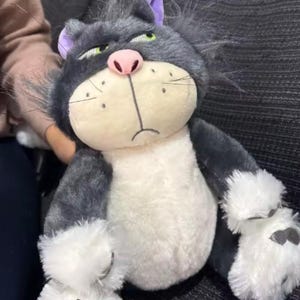 Puede incluir: Un peluche con forma de gato con una expresión de enfado. El gato es principalmente gris con detalles blancos en el pecho y las patas. Tiene una nariz rosa, ojos verdes y orejas moradas. Juguete para niños.