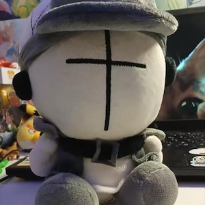 May include: A plush doll with a white body, gray hat, and gray accents. The doll has a black cross on its face and black ear coverings. It is wearing a gray vest and has gray, ball-shaped feet.