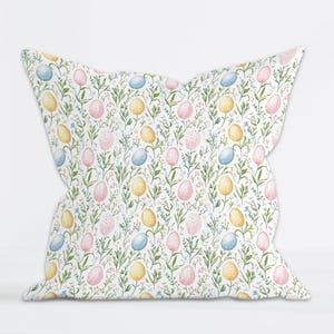 Peut inclure: Un coussin décoratif avec un motif répété d'œufs de Pâques aux couleurs pastel sur fond blanc. Les œufs sont roses, jaunes et bleus, avec des tiges et des feuilles vertes. Le coussin a une forme unique.