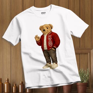 Puede incluir: Camiseta blanca con un oso de dibujos animados que lleva una chaqueta universitaria roja con las letras K, A y Ψ. El oso también lleva pantalones marrones y zapatillas blancas. La camiseta tiene el logo de Polo.