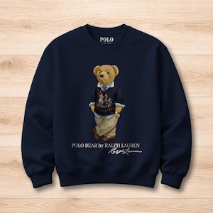 Puede incluir: Sudadera azul marino con un diseño de Polo Bear. El oso lleva un suéter, una camisa y pantalones color caqui. El texto "POLO BEAR by RALPH LAUREN" y la firma "Ralph Lauren" están impresos en la parte inferior.