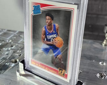May include: A graded basketball card of Shai Gilgeous-Alexander in a Los Angeles Clippers uniform, encased in a clear plastic holder. The card is displayed in a silver metal stand. The card is labelled "Rated Rookie" and includes the text "Clippers".