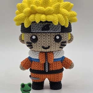 May include: A handcrafted figure made of small rubber bands featuring a character with spiky yellow hair, a black headband with a grey plate, and an orange and blue outfit. A small green frog figure stands next to the character on the ground.