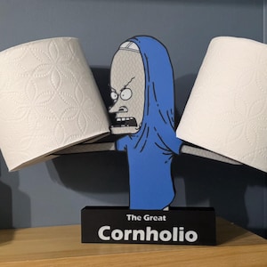 May include: A novelty toilet paper holder featuring a cartoon character in a blue robe, holding two rolls of white toilet paper. The base is black with the text "The Great Cornholio". The holder is on a wooden surface against a blue wall.