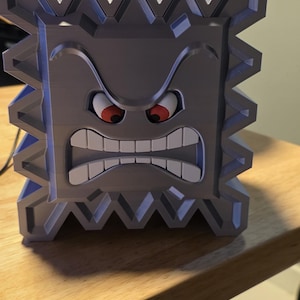 May include: A gray, 3D-printed tissue box shaped like a Thwomp from the Super Mario video game series. The box features an angry face with red eyes and white teeth, and is filled with white tissues.