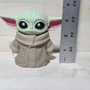 May include: A crocheted figure of a character with large black eyes, green skin, and large ears with pink inner ears. The figure is wearing a tan-colored robe. A ruler is next to the figure, measuring approximately 4 inches in height.