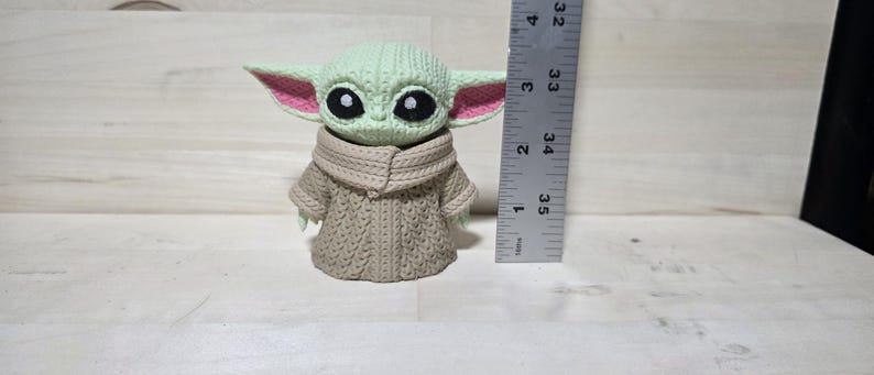 May include: A crocheted figure of a character with large black eyes, green skin, and large ears with pink inner ears. The figure is wearing a tan-colored robe. A ruler is next to the figure, measuring approximately 4 inches in height.