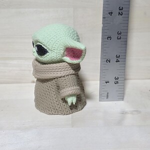 May include: A small, light green figurine of a character with large black eyes and pink-lined ears. The figure is wearing a tan-colored knitted coat and scarf. A ruler is positioned next to the figure, measuring approximately 3.5 inches in height.