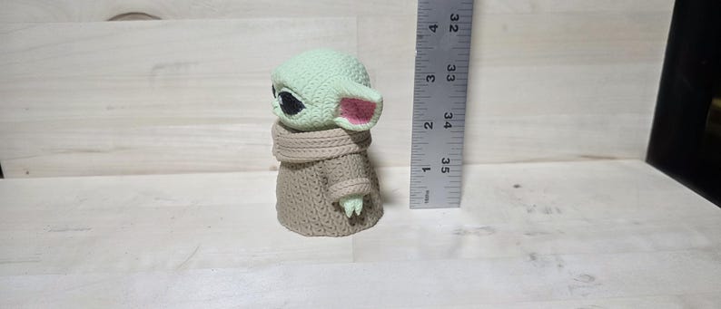 May include: A small, light green figurine of a character with large black eyes and pink-lined ears. The figure is wearing a tan-colored knitted coat and scarf. A ruler is positioned next to the figure, measuring approximately 3.5 inches in height.