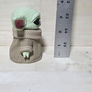 May include: A small, light green figurine of a character with large black eyes and pointed ears, wearing a tan robe. The figurine is approximately 3.5 inches tall. The character's head is light green with a red patch on each cheek.