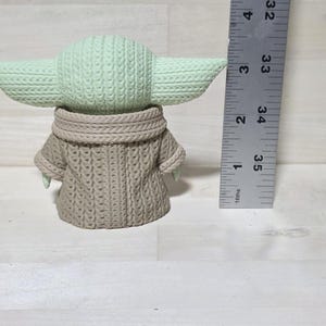 May include: A light green and tan crocheted figure of a character with large ears and a tan sweater. The figure is approximately 3.5 inches tall. The figure is on a light wood surface.