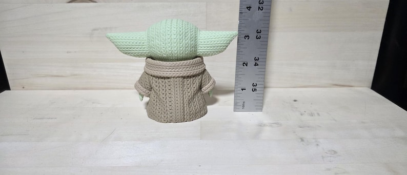 May include: A light green and tan crocheted figure of a character with large ears and a tan sweater. The figure is approximately 3.5 inches tall. The figure is on a light wood surface.