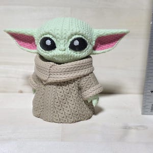 May include: A green and tan figurine of a character with large black eyes and large ears with pink inner ears. The figurine is wearing a tan sweater and a tan scarf. A ruler is next to the figurine.