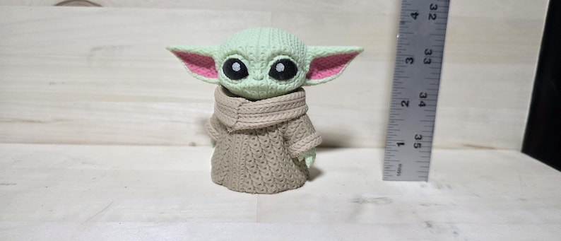 May include: A green and tan figurine of a character with large black eyes and large ears with pink inner ears. The figurine is wearing a tan sweater and a tan scarf. A ruler is next to the figurine.