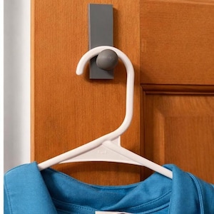 Könnte beinhalten: Ein weißer Türhaken mit einer grauen Montageplatte, der einen weißen Kleiderbügel hält. Ein blaues Hemd hängt an dem Kleiderbügel. Der Kleiderbügel ist an einer Holztür befestigt.